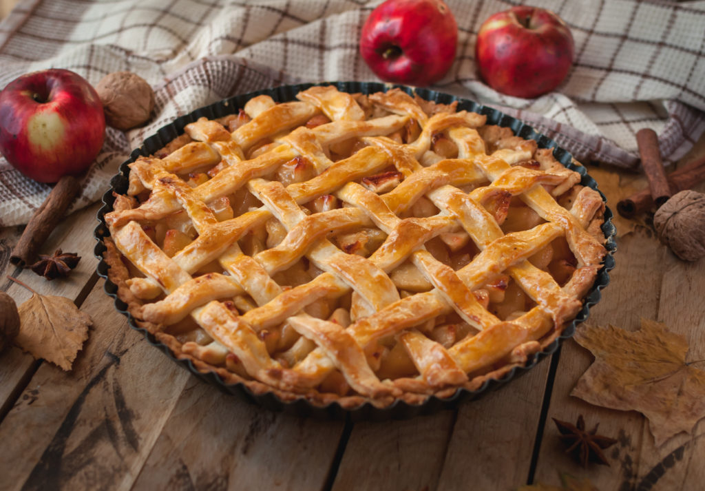 Image of apple pie on a table surrounded by apples, leaves, cinnamon sticks and walnuts.