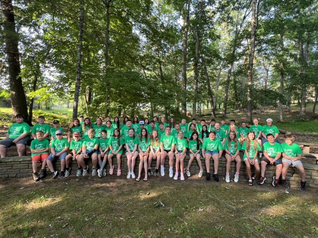 a large group of students in green T-shirts that say 4_H Camp posing in front of trees.
